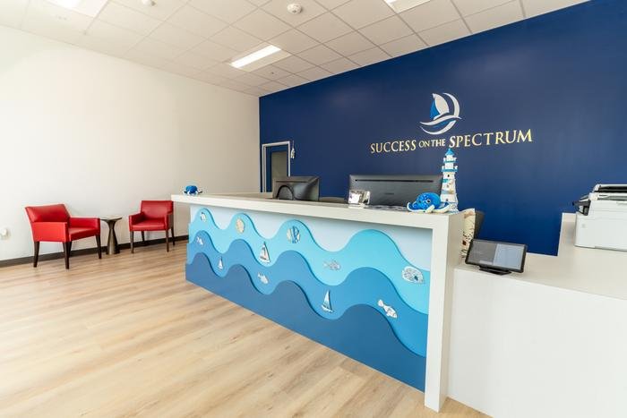 The SOS McKinney front desk and waiting area, with red armchairs and the Success On The Spectrum sailboat logo on the navy wall behind.