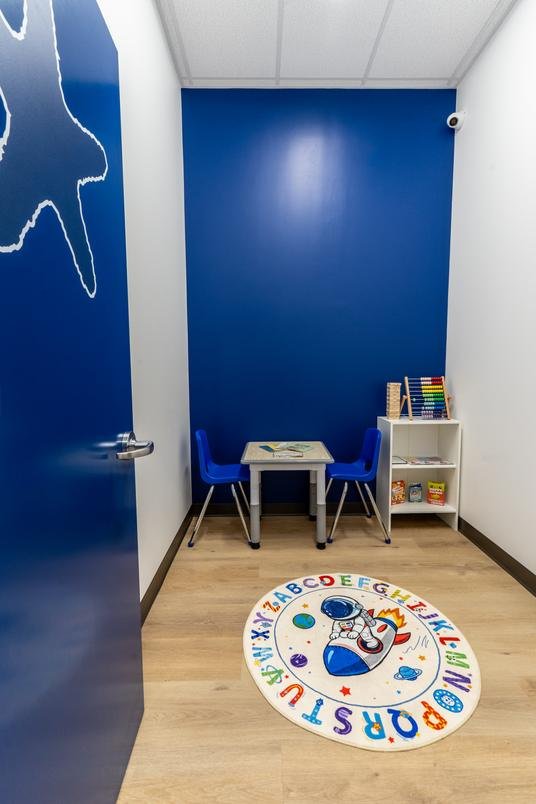 A quiet stateroom with a small table, two chairs, a shelf of games and an alphabet rug on the floor.